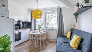 Uma cozinha moderna com armários brancos, uma mesa de jantar com cadeiras, uma luminária pendente amarela e um sofá cinza com almofadas amarelas.