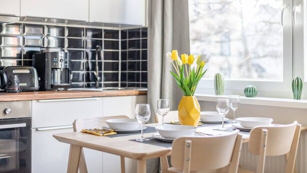 Uma cozinha moderna com área de jantar, apresentando um vaso amarelo com flores, uma janela com vista para árvores e um cacto no parapeito.