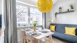 Uma área de refeições com uma mesa posta para quatro pessoas, um sofá com almofadas amarelas e uma janela com vista para edifícios.
