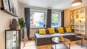 Ein modernes Wohnzimmer mit einem grauen Sofa, gelben Kissen, einem Glastisch und Blick auf die Stadt durch große Fenster.