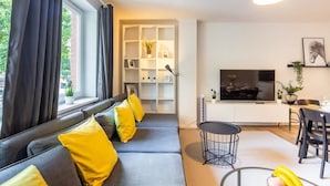 Ein modernes Wohnzimmer mit einem grauen Sofa, gelben Kissen, einem Essbereich mit einem schwarzen Tisch und Stühlen, einem Fernseher und einem Bücherregal.