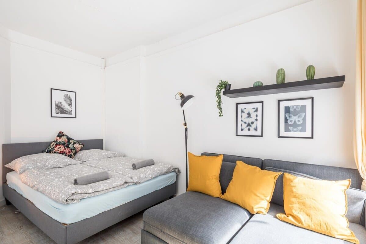Ein Schlafzimmer mit einem Bett, einem grauen Sofa und einem kleinen Tisch. An der Wand befinden sich gerahmte Bilder und ein Regal mit einer Pflanze.