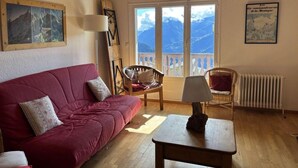 A cozy room with a wooden ceiling, a red sofa, a wooden table, and a view of mountains through large windows.