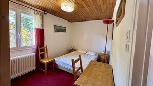 A small, cozy room with a wooden ceiling, a single bed, a wooden table, a chair, a window with curtains, and a radiator.