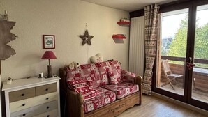 A cozy room with a wooden sofa, a white dresser, and a large window with a view of greenery. - 100m from resort center, 1st floor, balcony, tv, 25m², Alpe d'Huez (L'Alpe d'Huez)