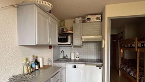 A compact kitchen with a microwave, toaster, and dishwasher. There is a small sink and a countertop with bottles. A bunk bed is visible in the background. - 100m from resort center, 1st floor, balcony, tv, 25m², Alpe d'Huez (L'Alpe d'Huez)