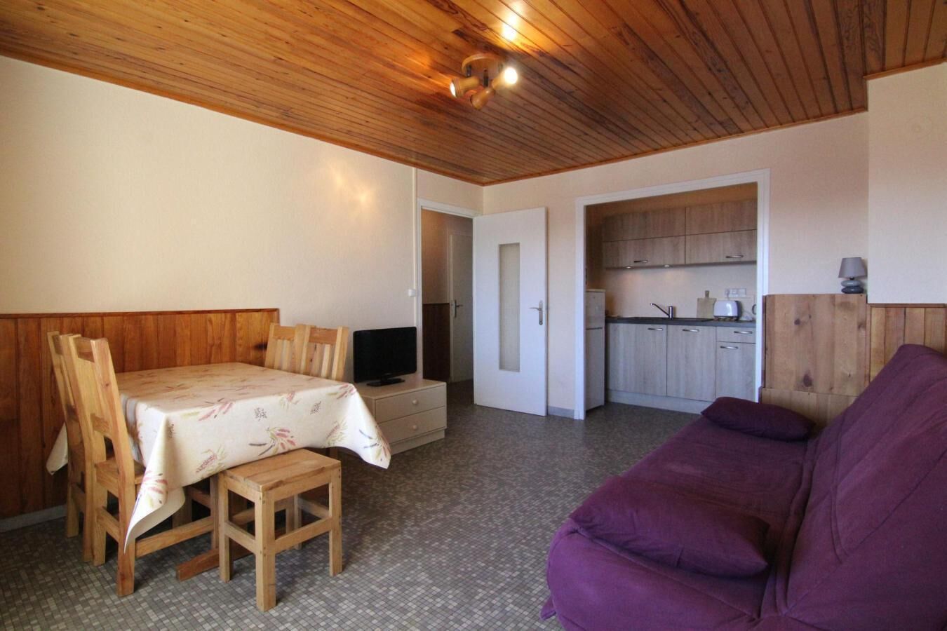 Una habitación con techo de madera, una cama con colcha estampada, un sofá púrpura, una pequeña mesa con una silla y una kitchenette al fondo.