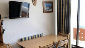 A dining area with a wooden table and chairs, a television mounted on the wall, and a window with curtains. - 80m from shops, 4th floor, balcony, tv, ski locker, 23m², Alpe d'Huez (L'Alpe d'Huez)
