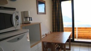 A kitchen with a microwave, coffee maker, and a refrigerator. There is a wooden table and a window with curtains. - 80m from shops, 4th floor, balcony, tv, ski locker, 23m², Alpe d'Huez (L'Alpe d'Huez)