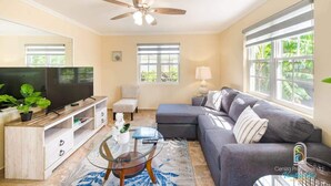A living room with a flat-screen TV, a ceiling fan, a grey sofa, a glass coffee table, and a large window with a view of greenery.