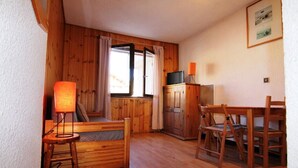 A cozy room with a wooden bed, a small table with a lamp, a TV on a cabinet, and a dining area with chairs.