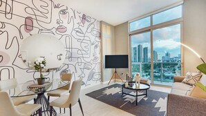 Un salon moderne avec vue sur la ville, une grande fresque murale abstraite et un coin repas avec une table en verre.