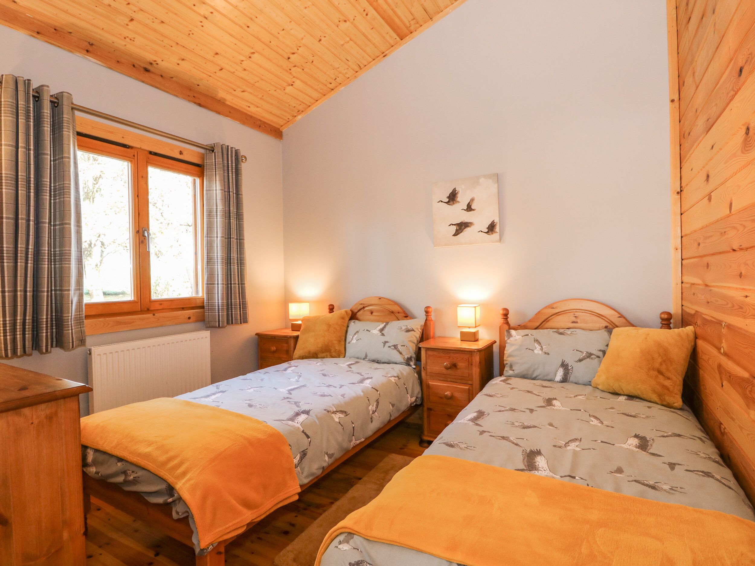 Ein gemütliches Schlafzimmer mit zwei Betten, Holz-Kopfteilen, einem Fenster mit Vorhängen, einer Nachttischlampe und einem Wandbild.