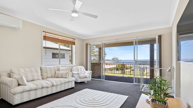 A spacious living room with a sofa, armchair, and a large sliding glass door leading to a balcony with a view of the sea. - Sunset on Loralyn by Book Jervis Bay (Saint Georges Basin)