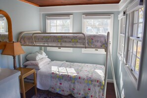 A room with a bunk bed, a bedside table with a lamp, and windows with white frames.