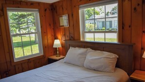 A wooden-paneled room with a bed, two windows, and a nightstand with a lamp. - 3 bedroom home with ac, walk to the beach! (West Dennis)