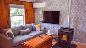 A living room with a gray sofa, a wooden coffee table, a black wood-burning stove, a white air conditioner, and a patterned wallpaper. - 3 bedroom home with ac, walk to the beach! (West Dennis)