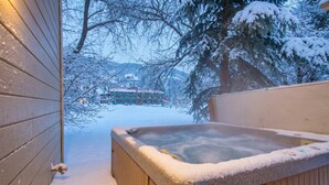 Un bain à remous en bois, avec de la neige sur le bord, un mur en bois et, en arrière-plan, une vue sur un bâtiment.