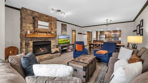 A living room with a stone fireplace, blue armchairs, a coffee table, and a television. - Modern 2BR Condo - Near Ski Lift; Walk Everywhere! (Park City)