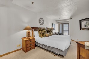 A bedroom with a bed, a nightstand, a lamp, a clock, and a window with blinds.