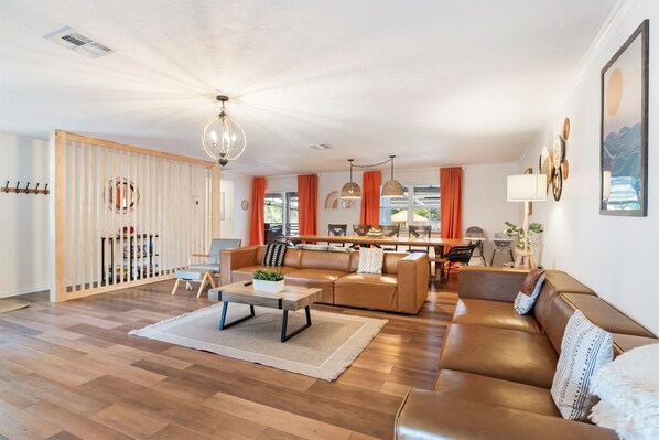 A spacious living room with a brown leather sofa, a wooden coffee table, and a dining area with orange curtains.