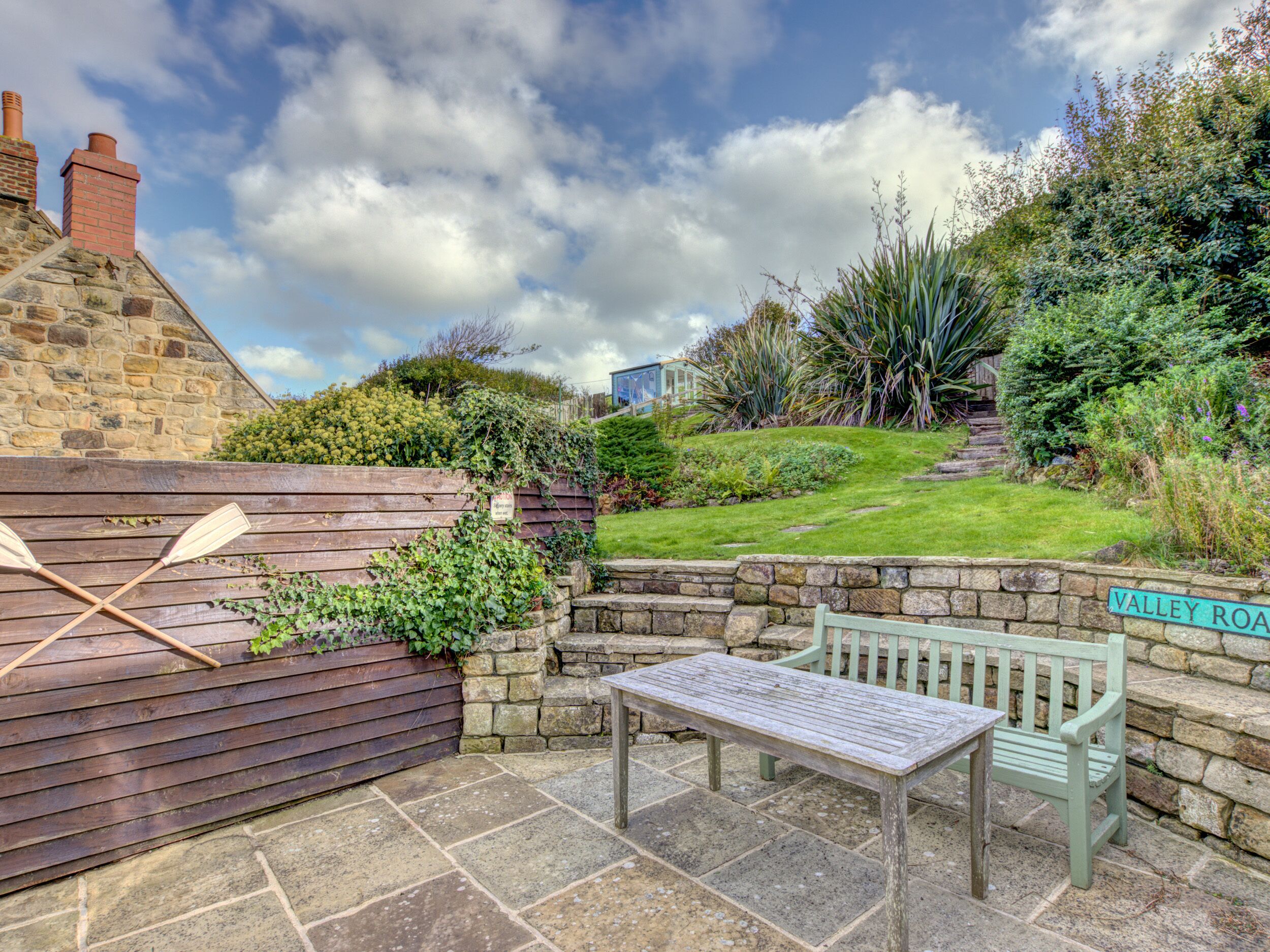 A stone patio with a wooden table and bench, surrounded by lush greenery and a stone wall.