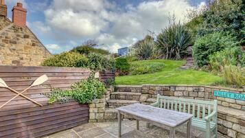 A stone patio with a wooden table and bench, surrounded by lush greenery and a stone wall.