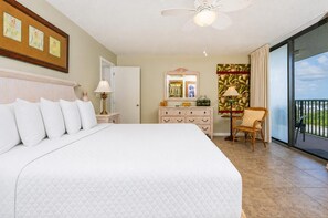 A hotel room with a large bed, a ceiling fan, a dresser, a chair, and a balcony with a view.