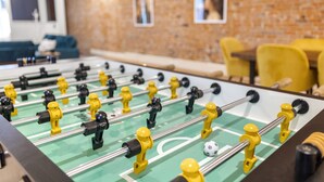 Une table de baby-foot avec un mur en briques et des chaises jaunes en arrière-plan.