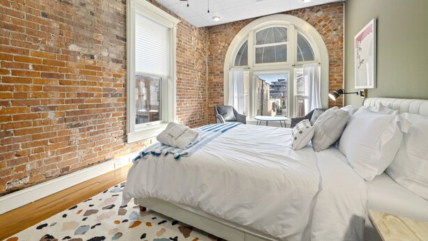 Une chambre avec un mur en briques, un grand lit, un ventilateur de plafond et une fenêtre donnant sur des immeubles.