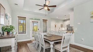A dining area with a wooden table and white chairs, a ceiling fan, and a kitchen in the background. - Pawsitively Paradise: Dog-friendly Home with Fenced Yard Close to Beach, Shops a (Oak Island)