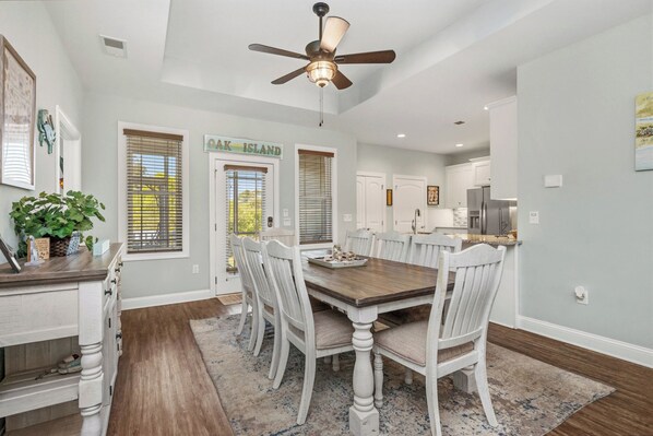A dining area with a wooden table and white chairs, a ceiling fan, and a kitchen in the background.