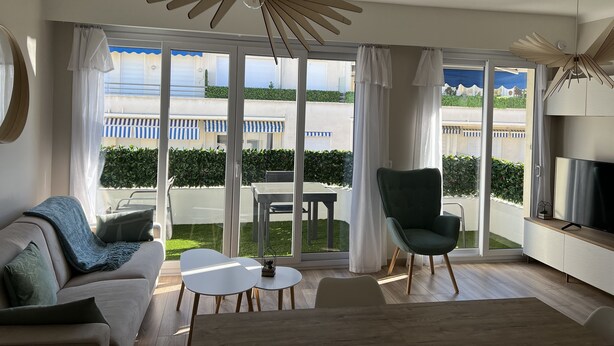 En moderne stue med sofa, stol, lille bord og fjernsyn. Store vinduer giver udsigt til en bygning med altaner og swimmingpool.