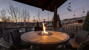 A round wooden table with a fire pit in the center, surrounded by wicker chairs, under a covered patio area with a view of a ski lift and mountains in the background. - CP2450 | Breathtaking Views | Ski In/Out | Winter Shuttle (Steamboat Springs)