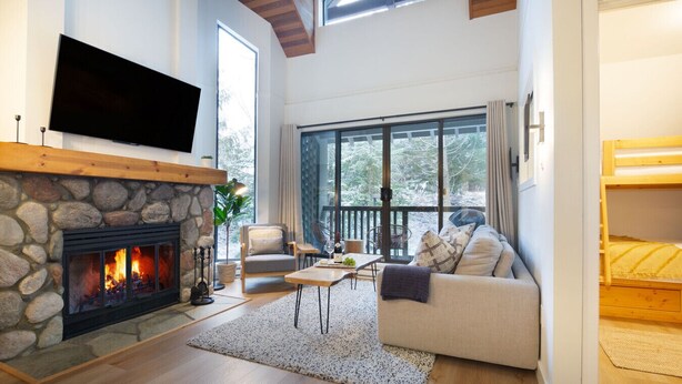Un salon chaleureux avec une cheminée en pierre, un téléviseur à écran plat fixé au mur, un canapé confortable et un balcon offrant une vue sur les arbres.