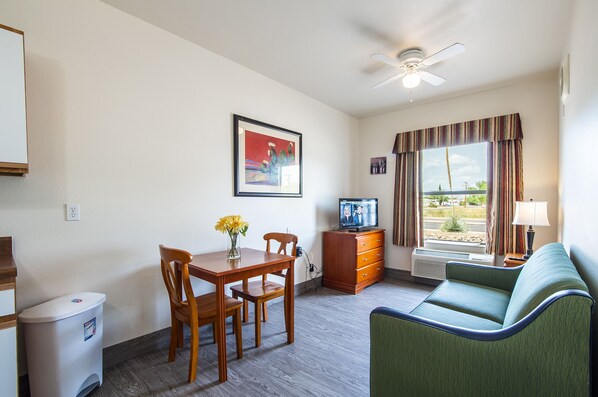 A hotel room with a dining table, chairs, a sofa, a TV, a ceiling fan, a window with curtains, and a wall-mounted air conditioner. - The Vistas (Sierra Vista)