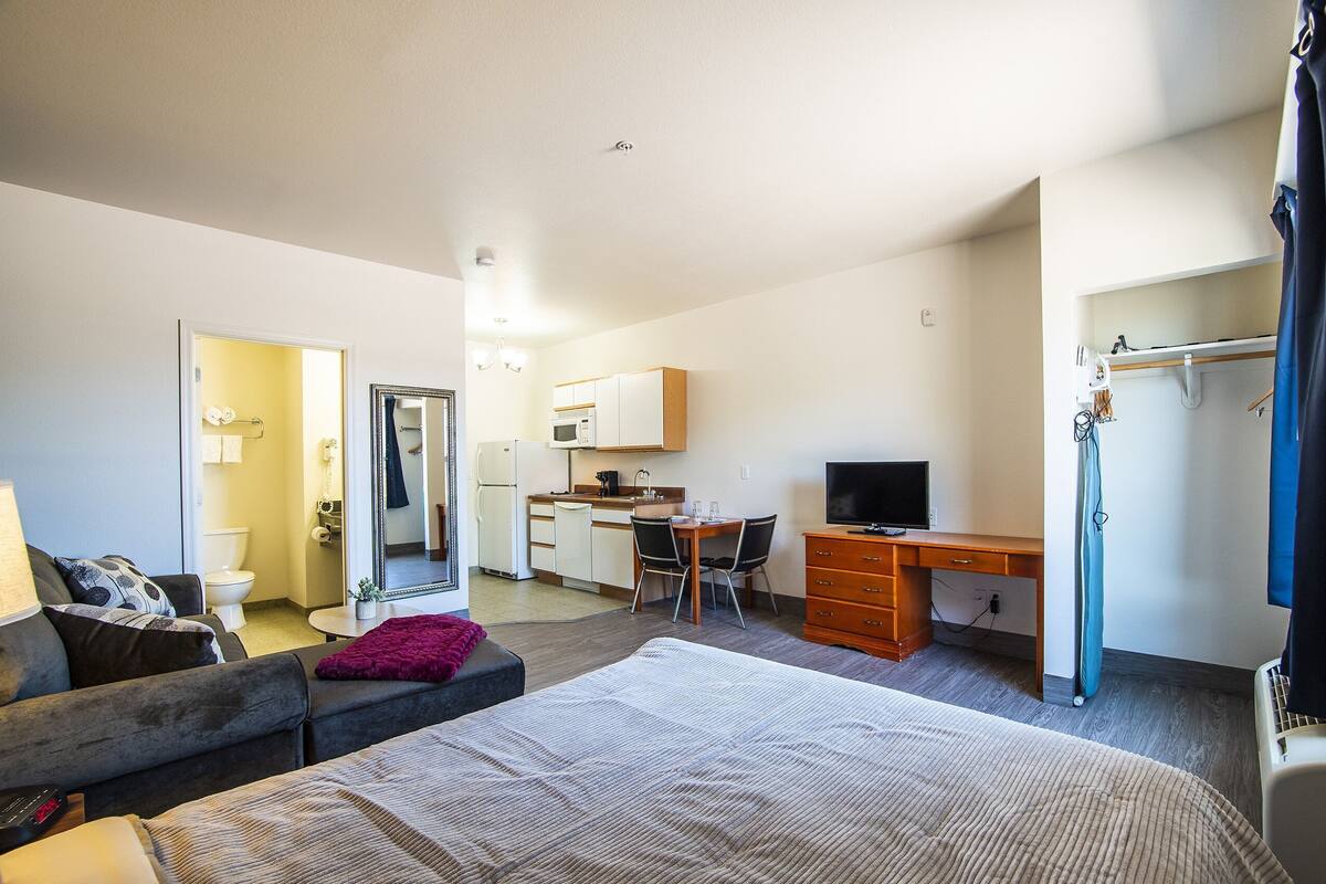 Een hotelkamer met een bed, bureau, stoel, televisie, spiegel, kitchenette en een badkamer.