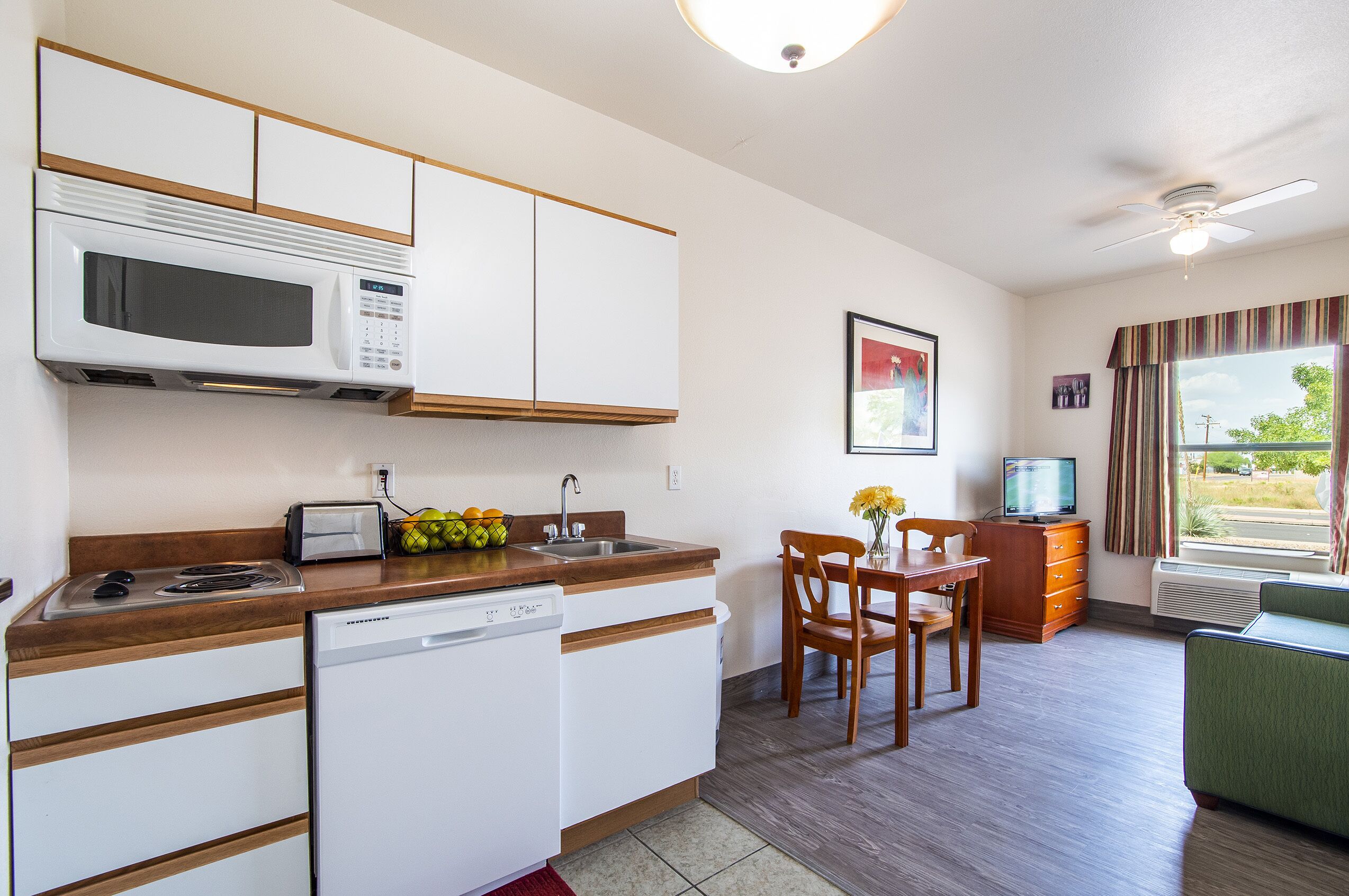 A compact kitchen with a microwave, toaster, and dishwasher. There is a dining area with a table and chairs, and a television on a stand.