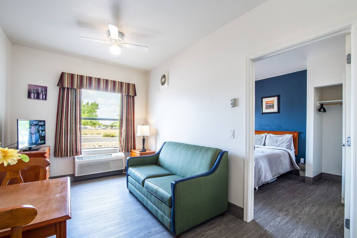 Een hotelkamer met een groene bank, een bed met een blauw hoofdeinde, een eettafel met stoelen, een televisie en een raam met gordijnen.