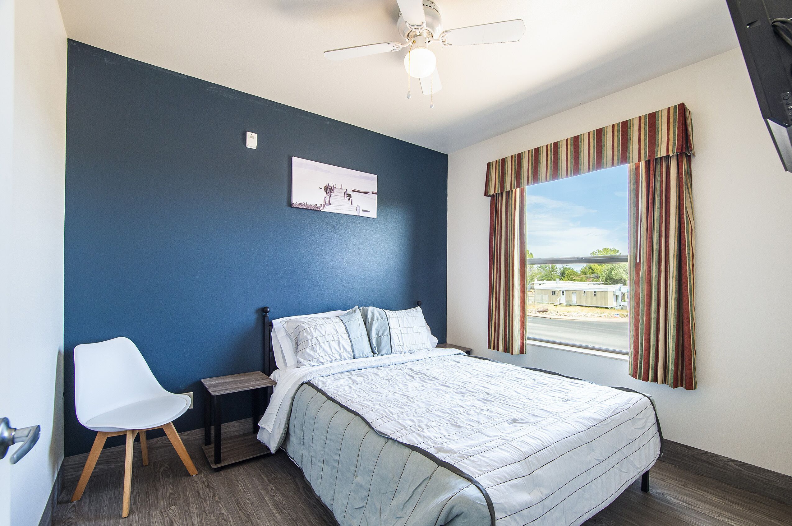 A bedroom with a bed, a chair, a nightstand, a ceiling fan, a window with curtains, and a picture on the wall.