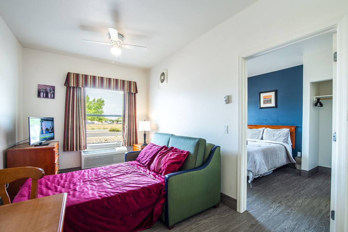 Een hotelkamer met een bed, een bureau met een televisie, een raam met gordijnen en een plafondventilator.