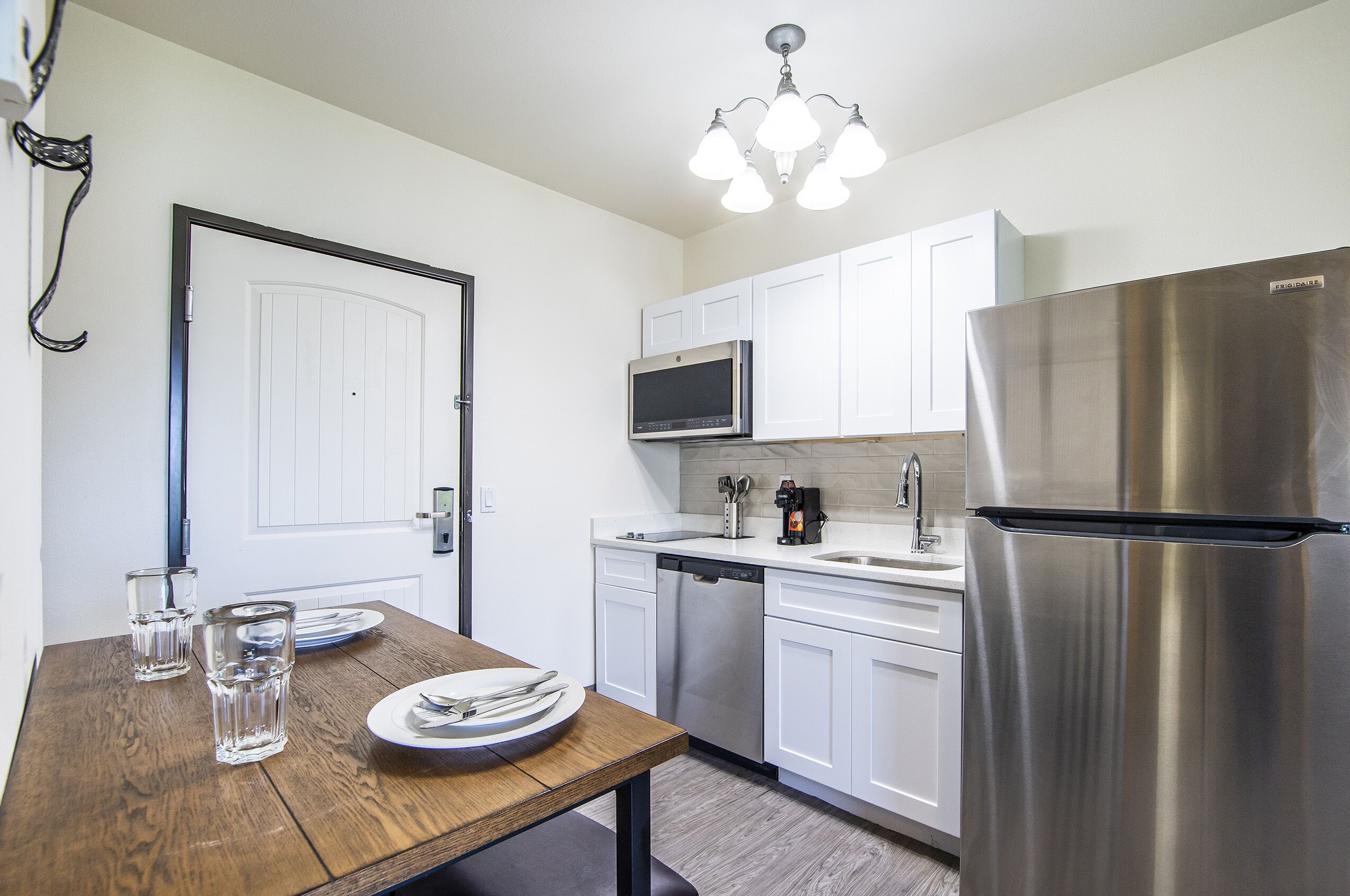 A modern kitchen with white cabinets, stainless steel appliances, a wooden dining table with plates and glasses, and a chandelier.