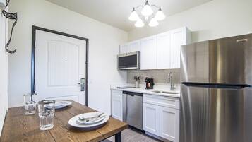 A modern kitchen with white cabinets, stainless steel appliances, a wooden dining table with plates and glasses, and a chandelier.