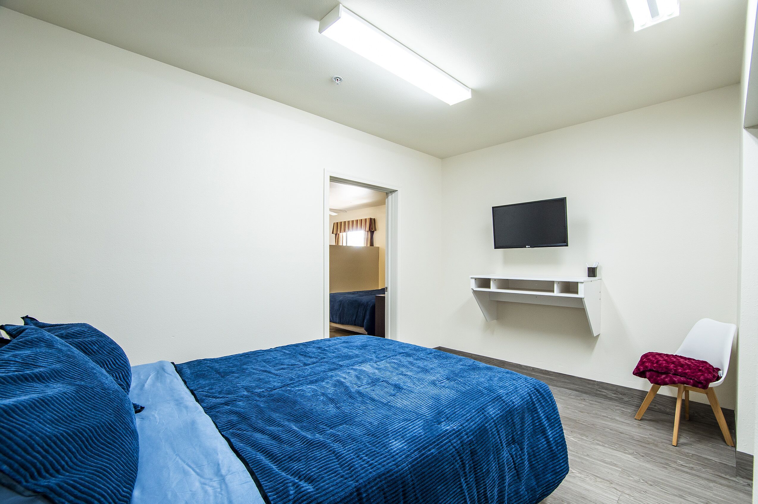 A hotel room with a bed, a TV mounted on the wall, a chair with a blanket, and a small shelf.