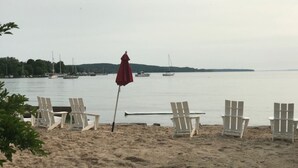 Une plage avec des chaises blanches et un parasol rouge, des bateaux à l’arrière-plan et une mer calme.