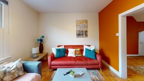 A living room with a red sofa, a small table, a lamp, and a painting on the wall. - Mill House | Oxford Holiday Let | 2 Beds & Sleeps 4 (Oxford)
