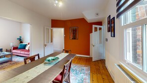 A dining area with a long table, chairs, and a rug. There is a sofa and a lamp in the adjacent room. - Mill House | Oxford Holiday Let | 2 Beds & Sleeps 4 (Oxford)
