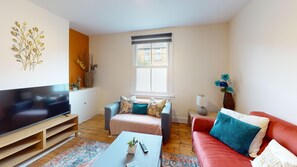 A living room with a flat-screen TV, a sofa, a red couch, a coffee table, and a window with a view of buildings. - Mill House | Oxford Holiday Let | 2 Beds & Sleeps 4 (Oxford)