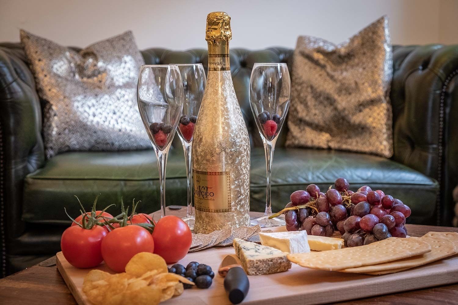 Una mesa con una botella de champán, copas, uvas, queso, tomates y galletas.