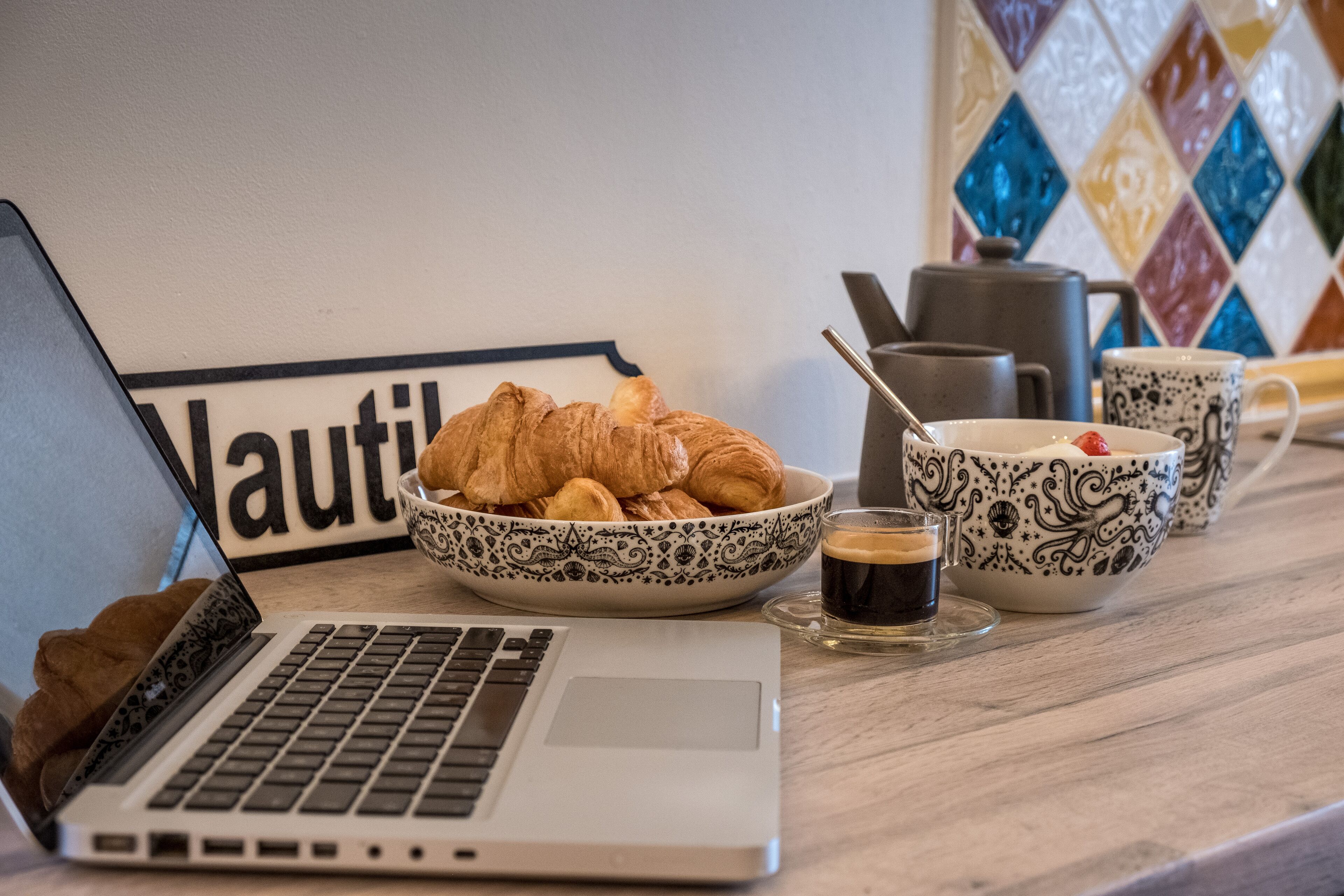 Um laptop, um prato de croissants, uma chávena de café e uma chaleira sobre uma mesa.